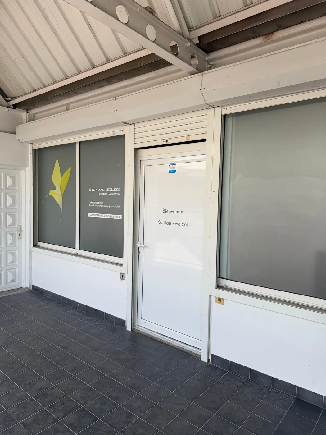 Entrée du cabinet Jéquece Nutrition : une porte blanche marquée « Bienvenue, Kontan vwè zòt » et une vitrine illustrée d’un oiseau jaune